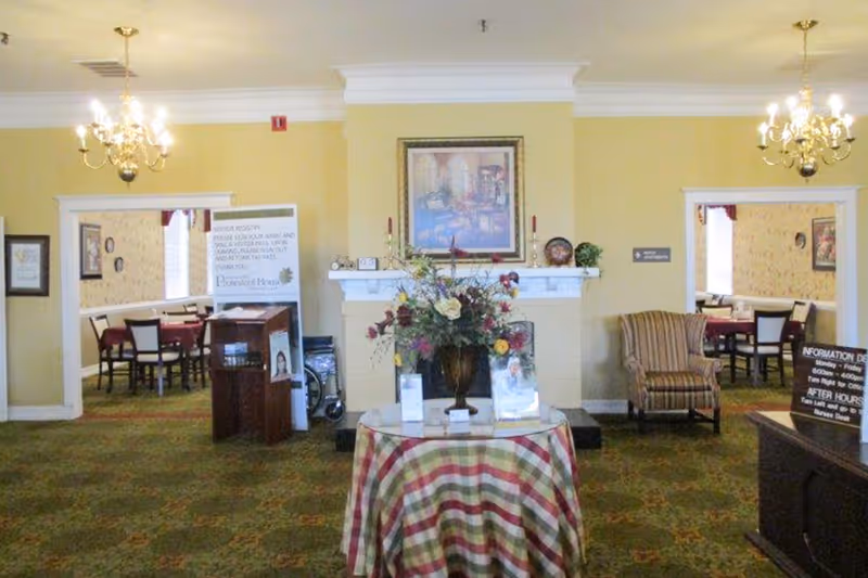 Interior view of a senior living facility lobby area with a round table covered with a checkered cloth and a large floral arrangement in the center. There are two chandeliers hanging from the ceiling, a fireplace with a framed painting above it, and two doorways leading to dining rooms with tables and chairs. An upholstered armchair is placed near the right doorway.