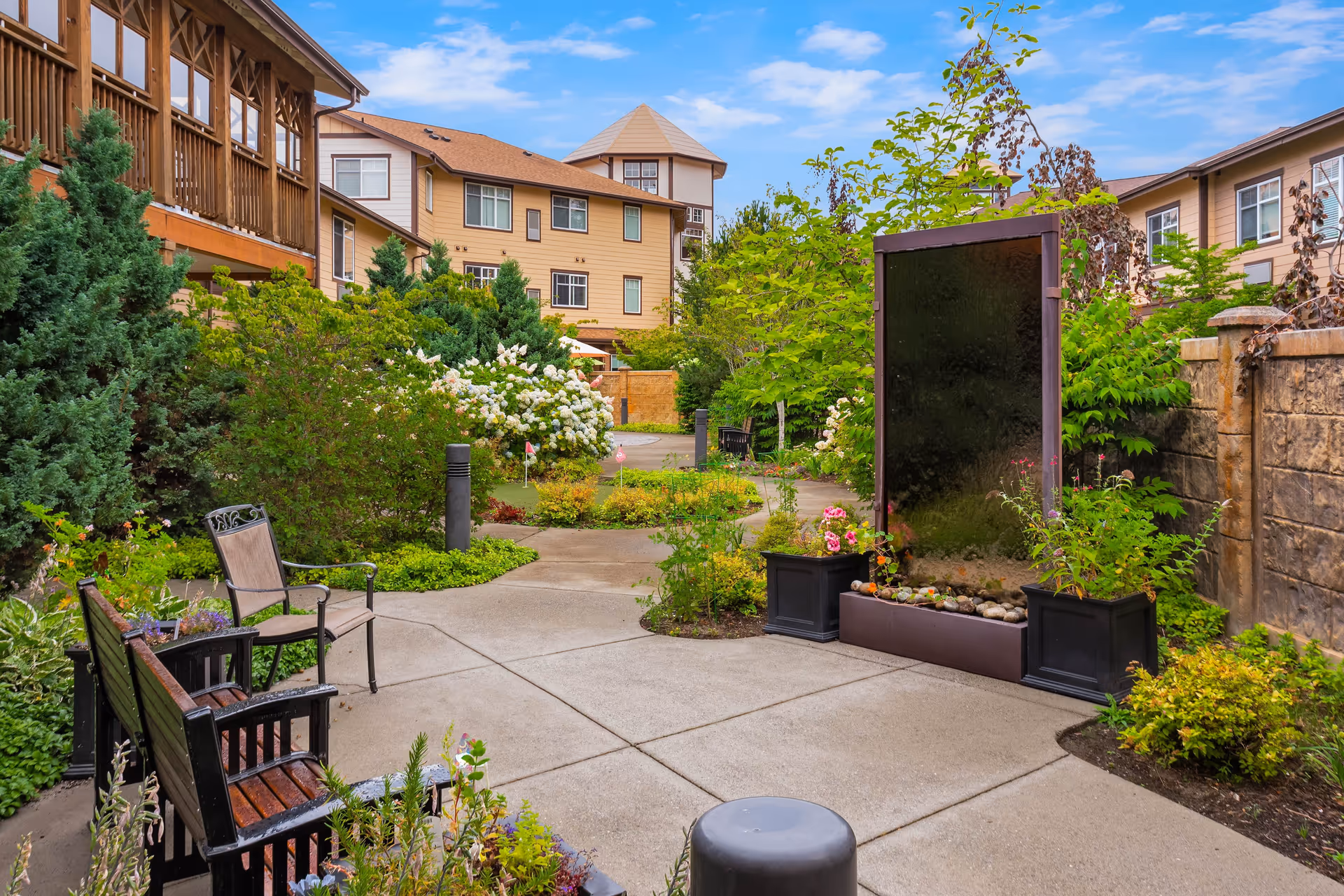 A landscaped outdoor garden area at Quail Park of Lynnwood featuring a paved walkway, benches, chairs, lush greenery, flowering plants, and a vertical water feature. Residential buildings with balconies and windows surround the garden under a partly cloudy blue sky.