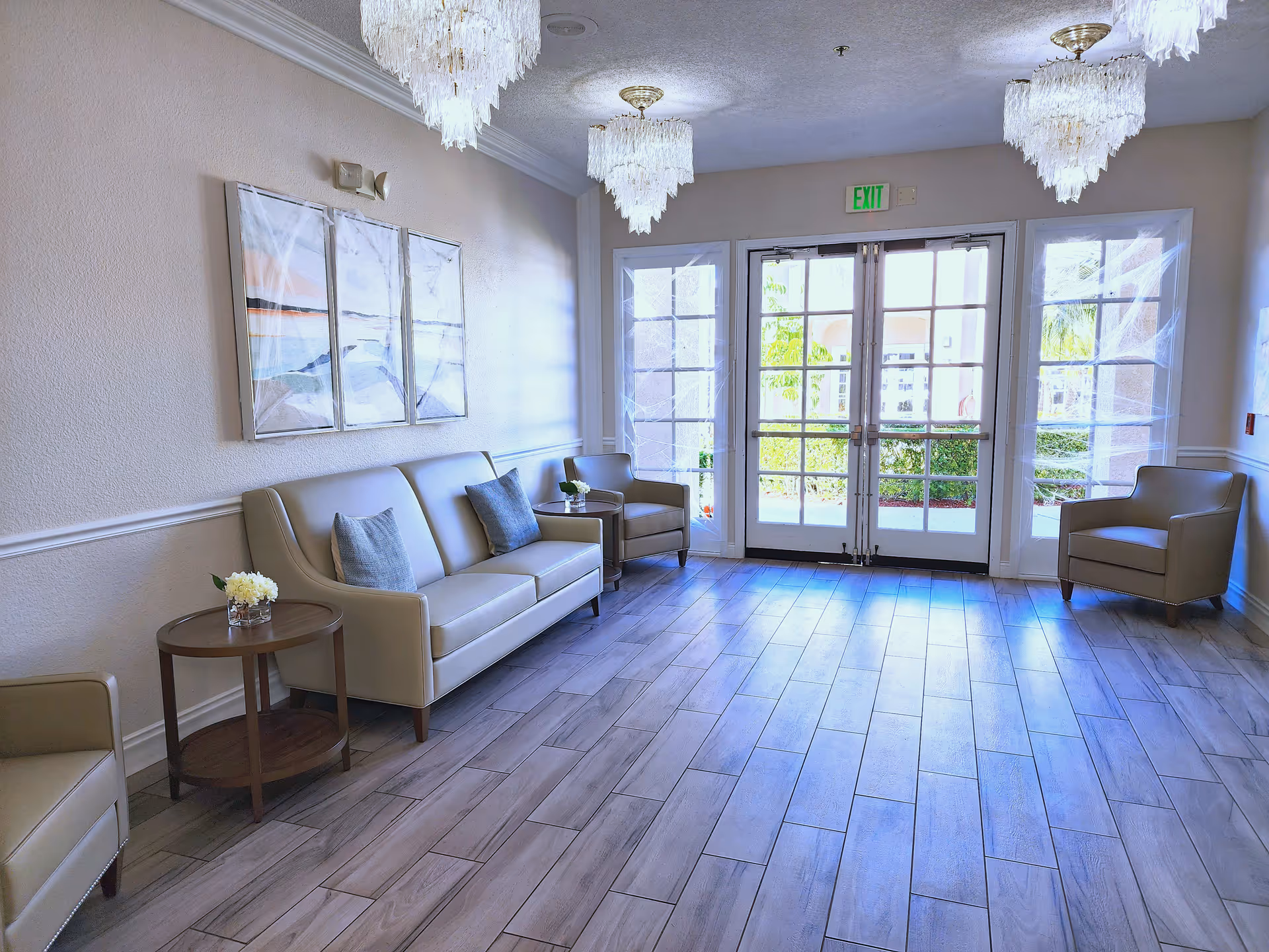 A bright and clean seating area with a beige leather sofa and matching armchairs arranged around small round wooden tables with flower vases. The room has large glass double doors letting in natural light, three elegant chandeliers hanging from the ceiling, and a triptych wall art above the sofa. The floor is covered with light wood-look tiles.