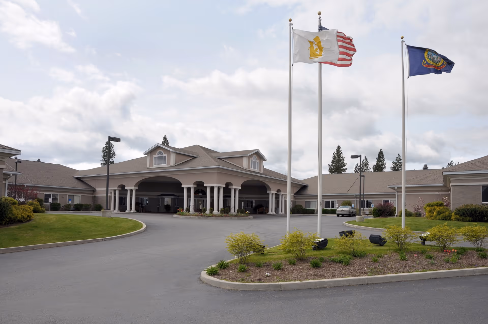 Front exterior view of the Life Care Center of Coeur d'Alene building with a covered entrance, three flagpoles displaying flags, a circular driveway, and landscaped greenery under a cloudy sky.