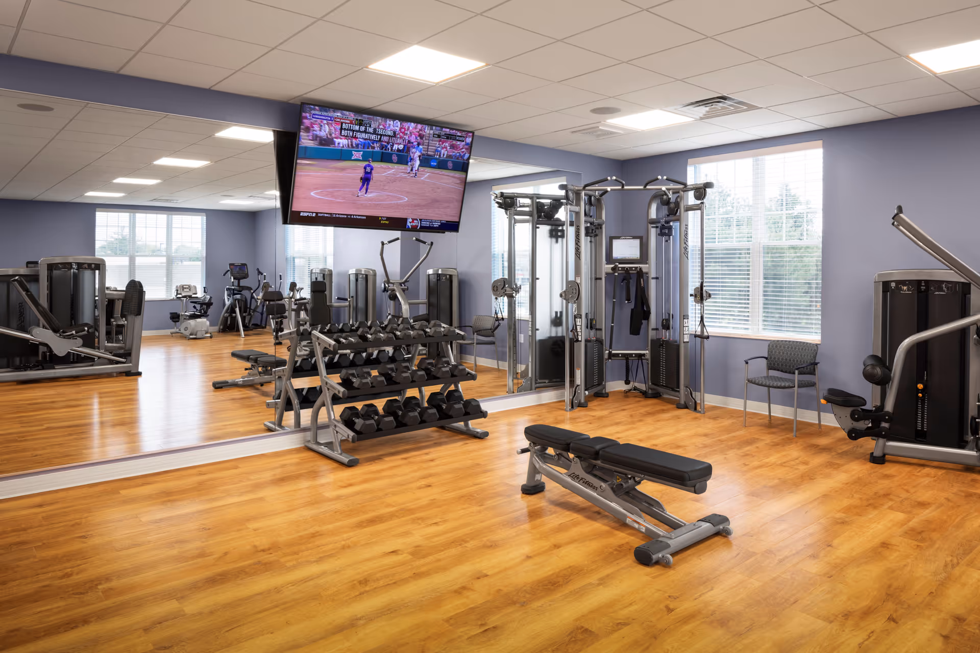 A well-lit fitness room with wooden flooring, various exercise machines, a rack of dumbbells, a bench, and a large wall-mounted TV showing a sports game. The room has large windows with white blinds and a wall mirror reflecting the equipment.