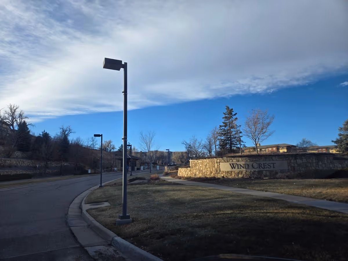 Stone entrance sign reading "Wind Crest" beside a curved road with lampposts and buildings under a blue sky.