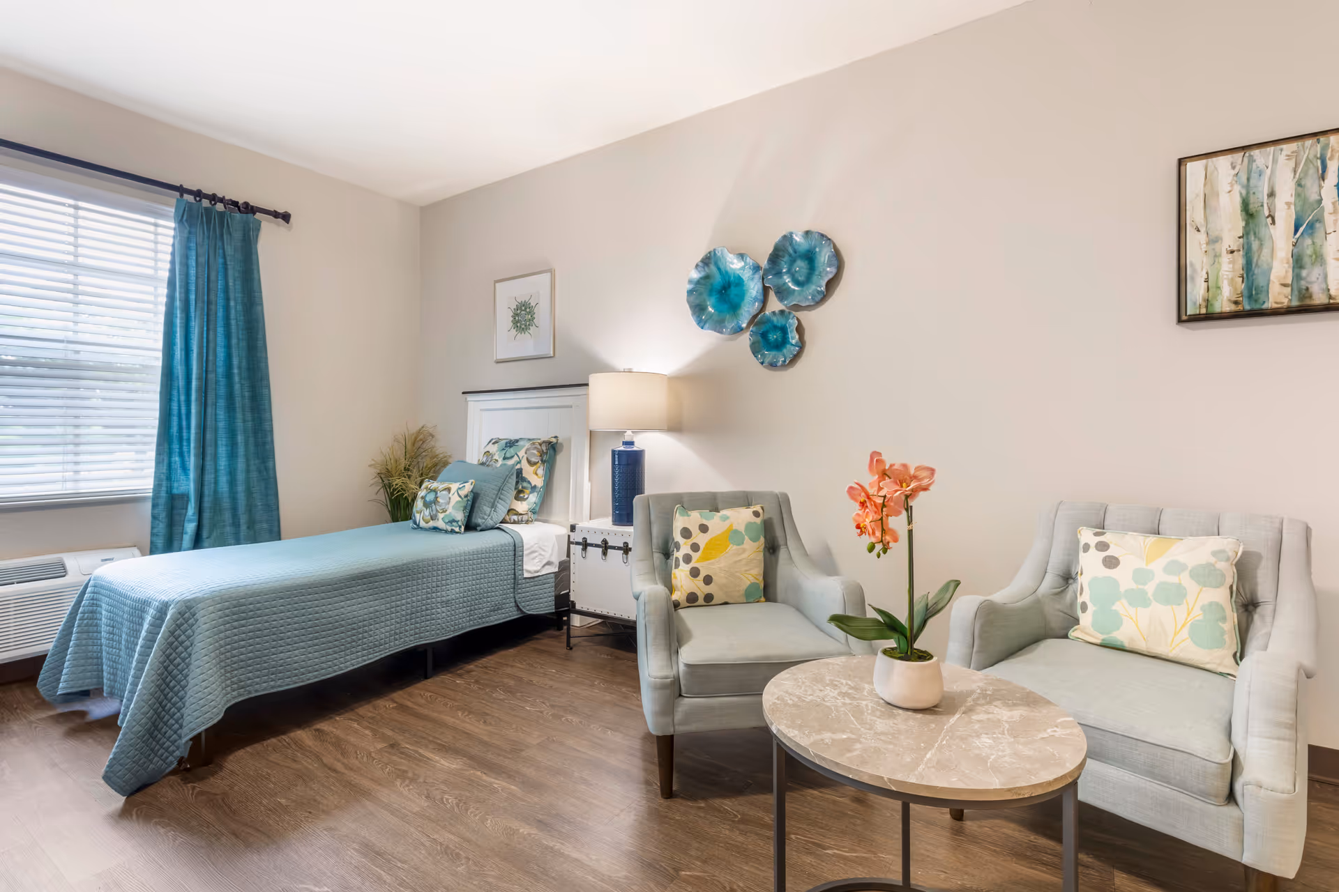 A bright and cozy bedroom with a single bed covered in a teal quilt and matching pillows. Next to the bed is a white nightstand with a blue lamp. There are two light blue armchairs with patterned cushions and a round marble-top table with a potted orchid in front of them. The room has wood flooring, a window with teal curtains, and decorative wall art including blue ceramic flowers and framed pictures.