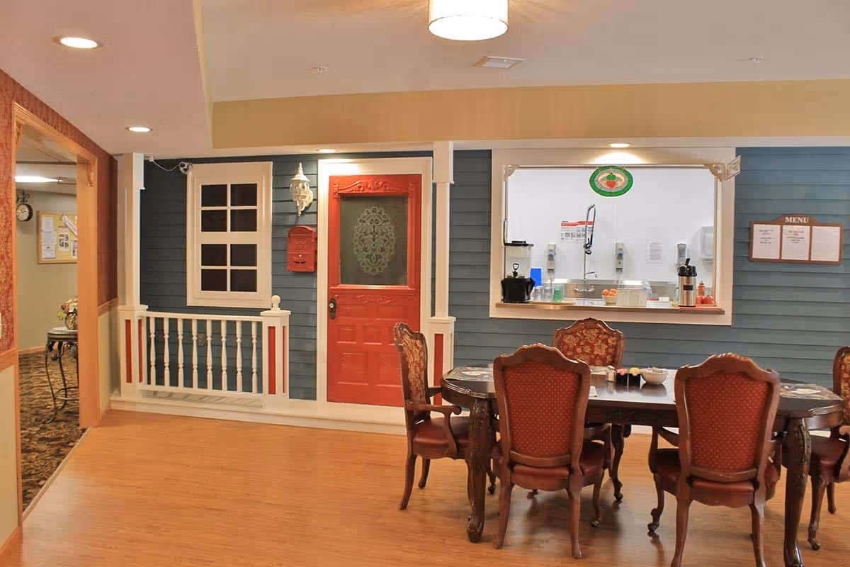 Interior view of a senior living facility dining area with a wooden table and six upholstered chairs. The wall features a decorative red door with a frosted glass panel, a window with white trim, a red mailbox, and a serving window to a kitchen area. The floor is wooden, and the ceiling has recessed lighting.