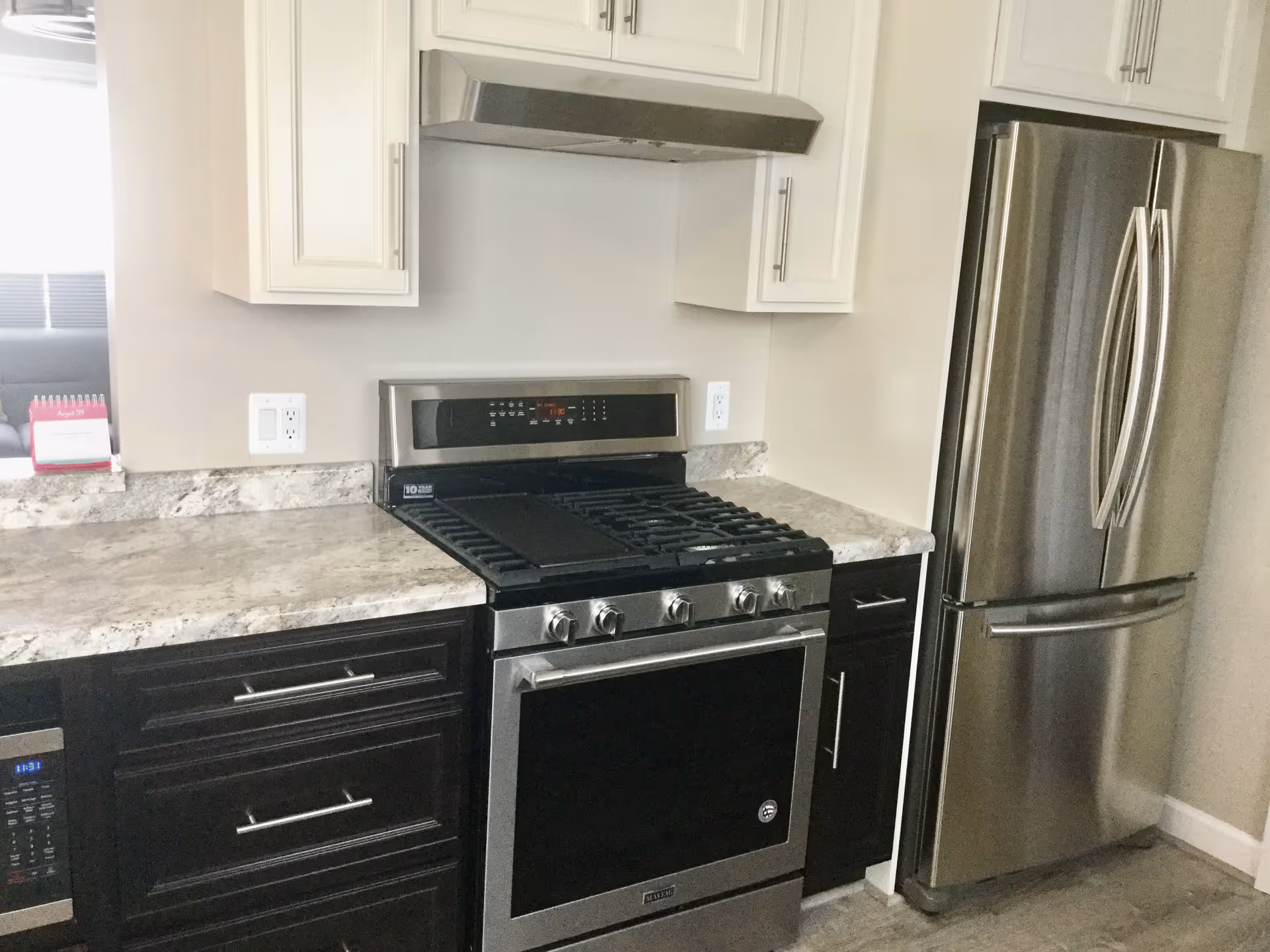 Modern kitchen with stainless steel appliances including a refrigerator and a gas stove with oven. The kitchen has white upper cabinets and dark lower cabinets with silver handles, and a light-colored granite countertop.