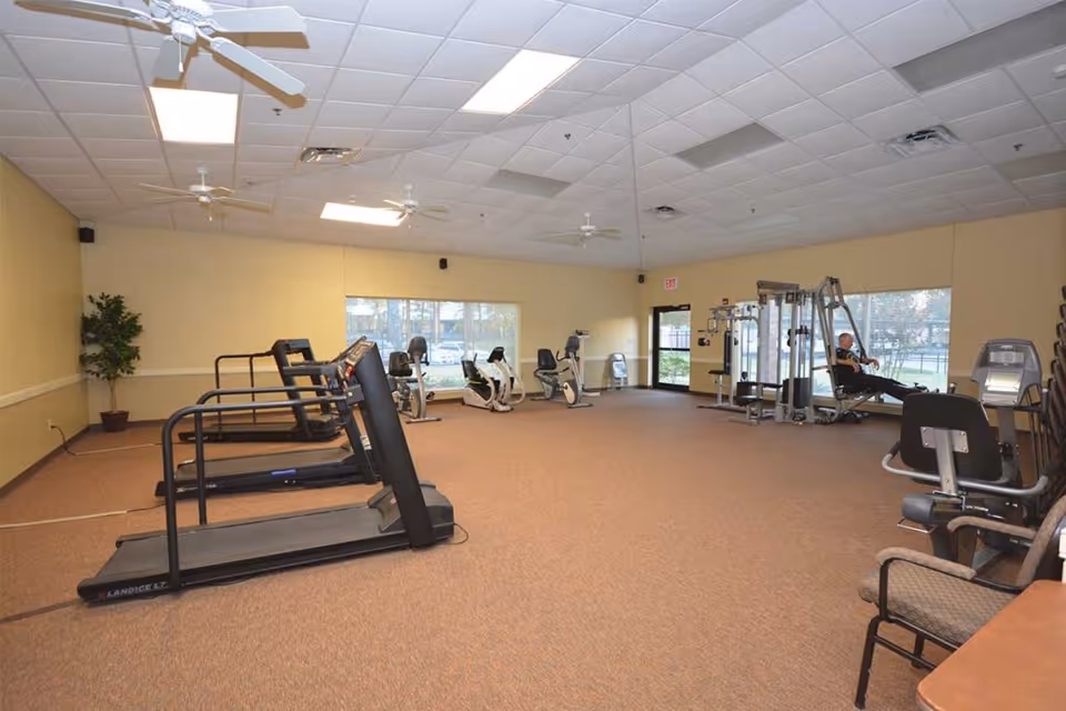 A spacious fitness room with several treadmills, stationary bikes, and weight machines. The room has beige walls, a carpeted floor, large windows letting in natural light, ceiling fans, and a potted plant in the corner. A person is using one of the weight machines near the windows.