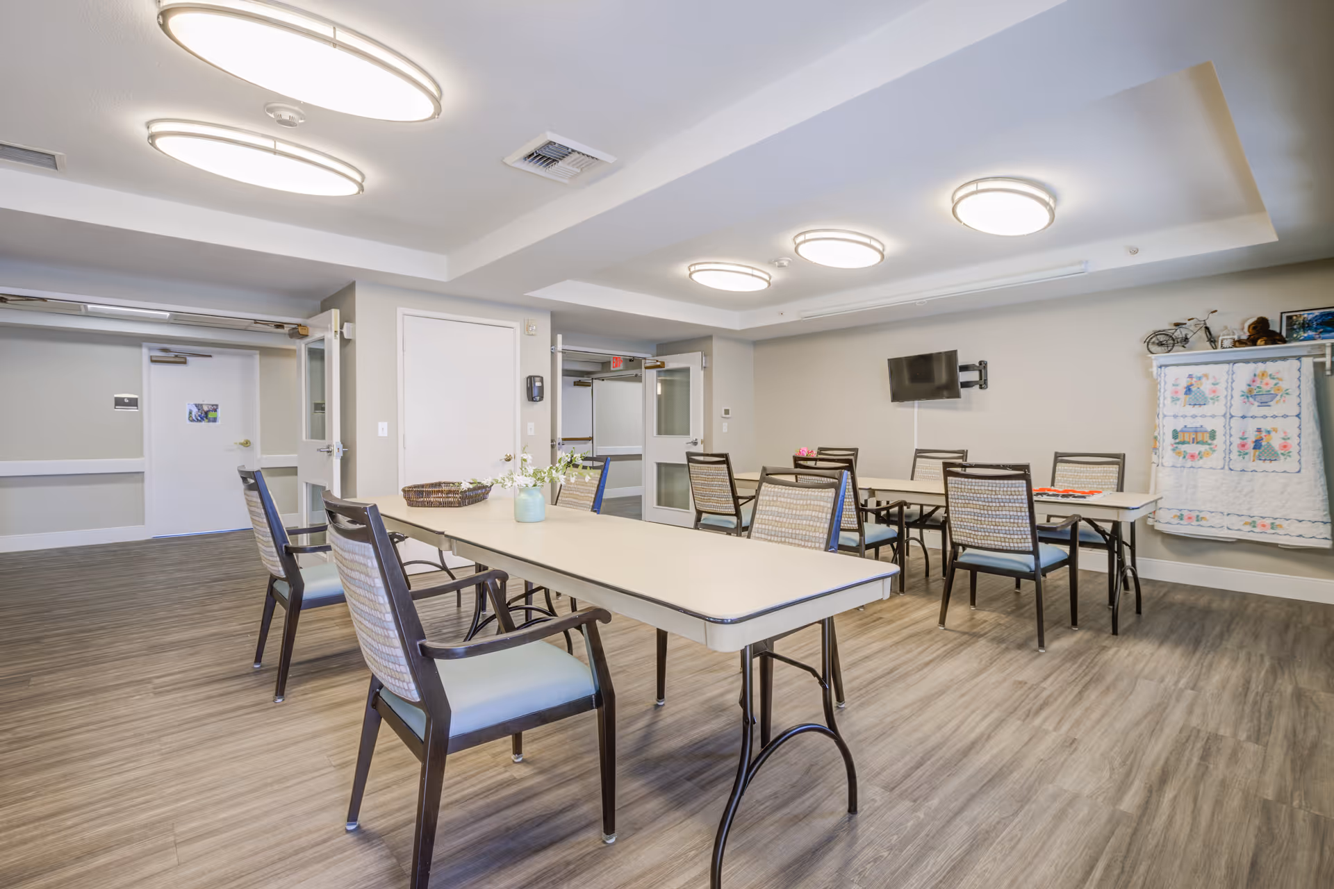 A well-lit common room with two rectangular tables and several chairs arranged around them. The room has light-colored walls, wood-patterned flooring, and modern circular ceiling lights. A small TV is mounted on the wall, and a quilt hangs on a rack in the corner. Double doors and a white door are visible in the background.