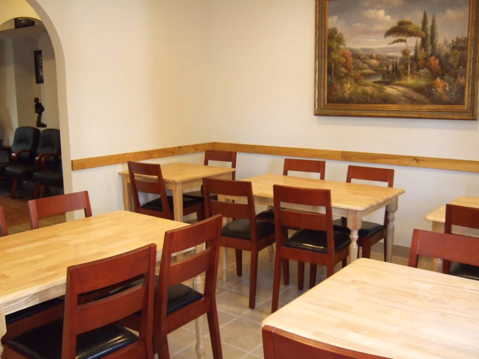 A dining area with several wooden tables and chairs with dark cushions. The walls are light-colored with a wooden trim, and there is a large framed painting of a landscape hanging on the wall. In the background, there is an archway leading to another room with additional seating.
