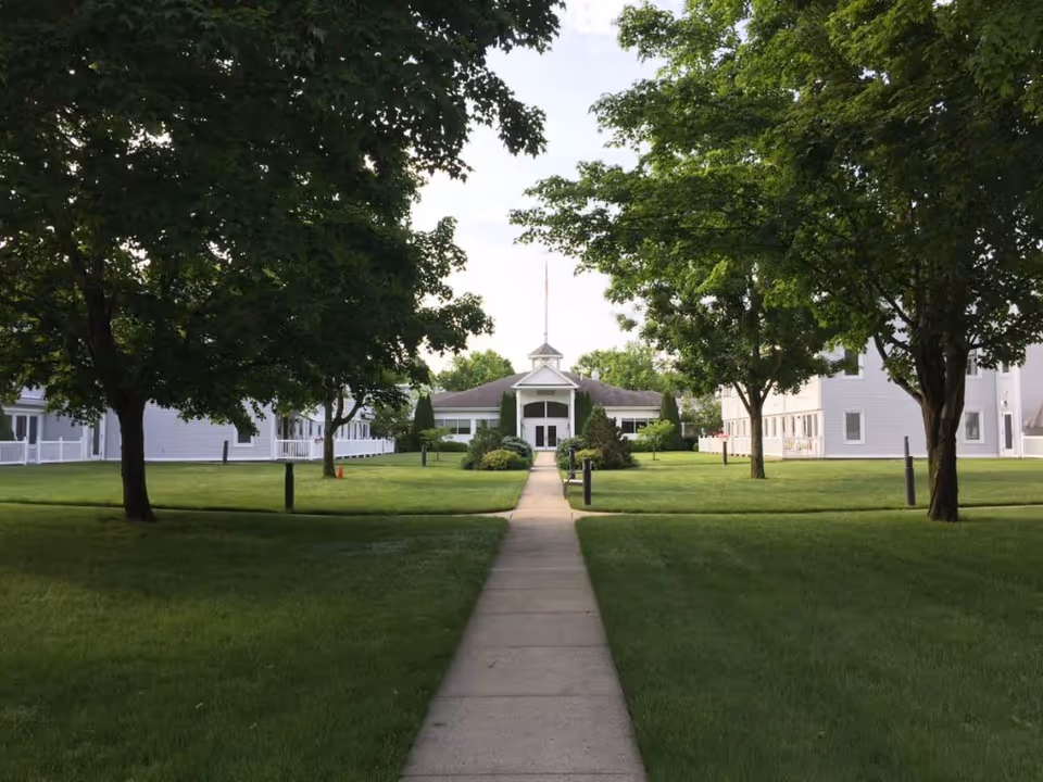 Concrete walkway lined by green lawns and trees leading to the entrance of a white senior living building.