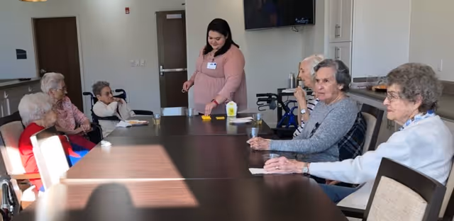 A group of elderly women sitting around a long table in a well-lit room, with a caregiver standing and interacting with them. The room has light-colored walls, a door, a television mounted on the wall, and cabinets in the background.