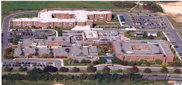 Aerial view of the Gurwin Jewish Nursing & Rehabilitation Center showing multiple connected brick buildings with flat roofs, surrounded by parking lots, roads, and landscaped green areas.