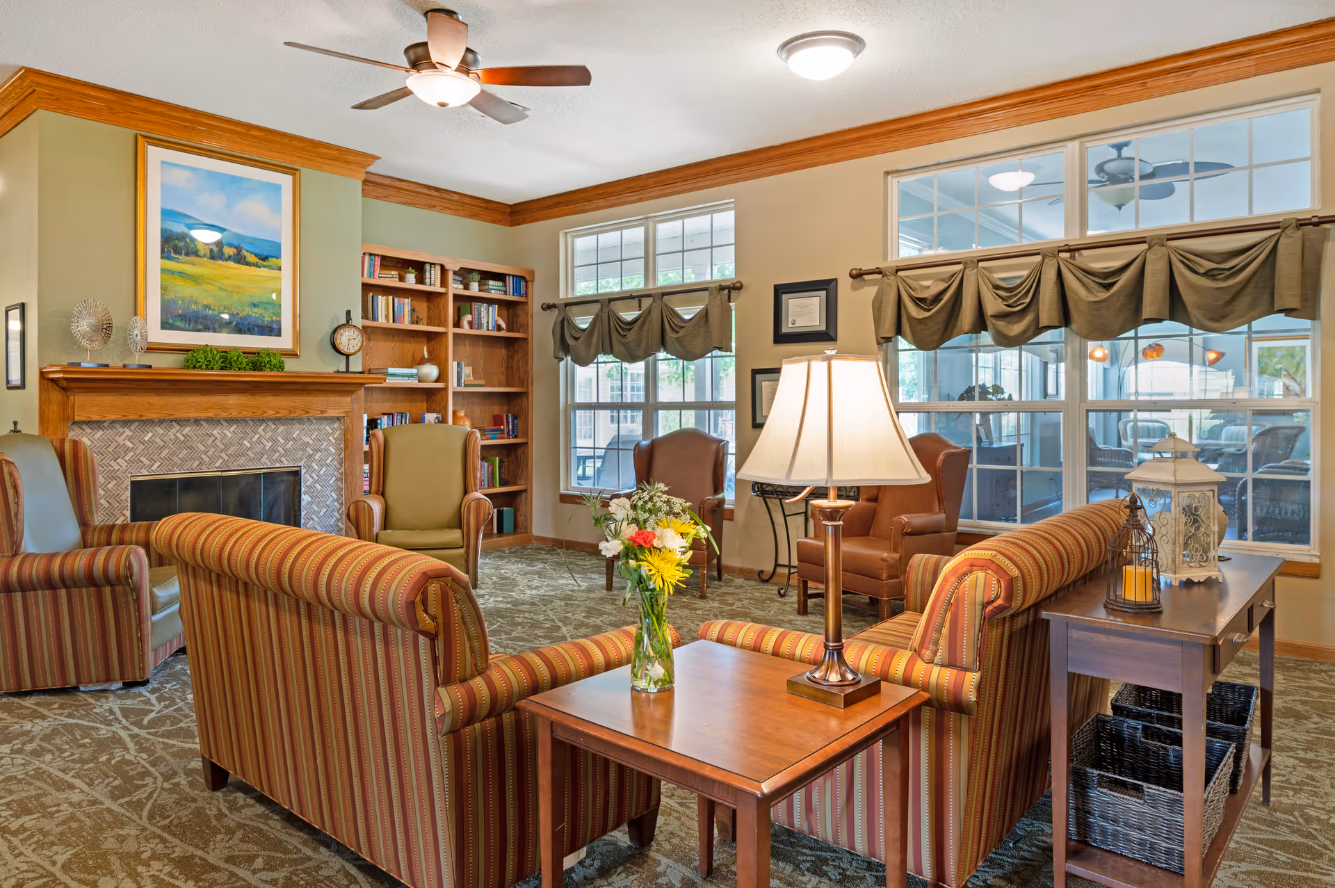 A cozy living room with striped sofas, armchairs, a wooden bookshelf, a fireplace with a framed landscape painting above it, a ceiling fan, and large windows with draped curtains letting in natural light. A wooden side table with a lamp and a vase of flowers is in the foreground.