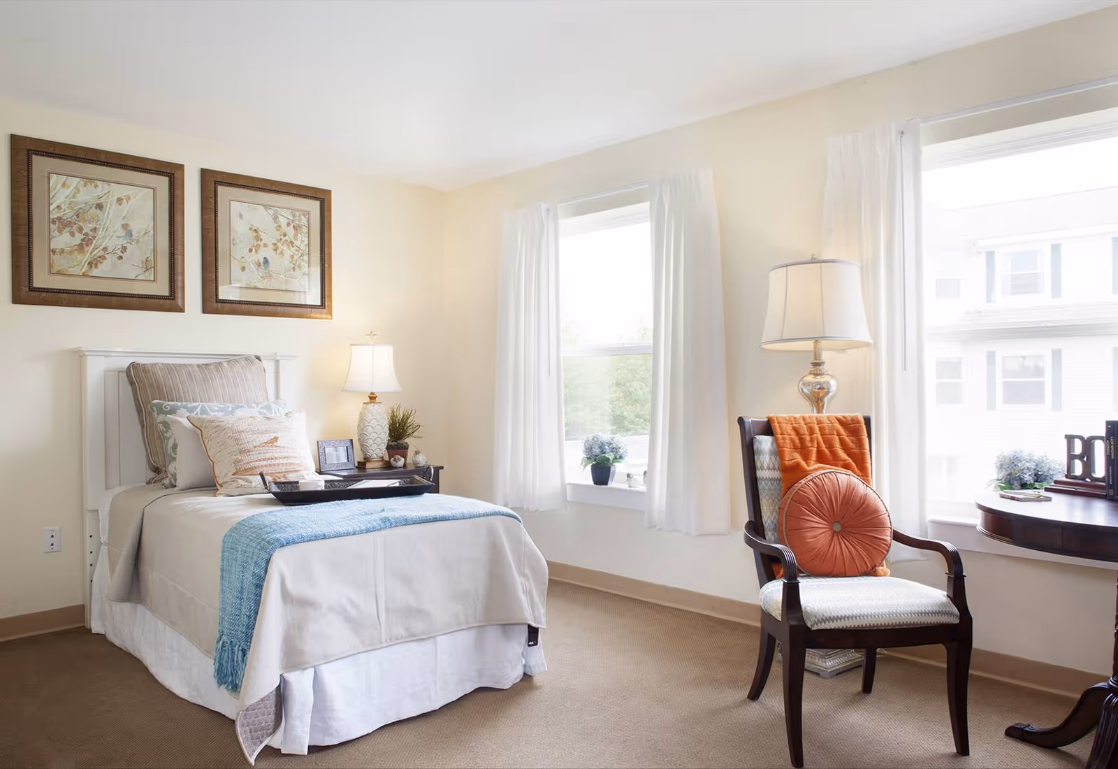 Bright, neatly staged bedroom with a single bed, bedside table and lamp, and an upholstered chair by two windows.
