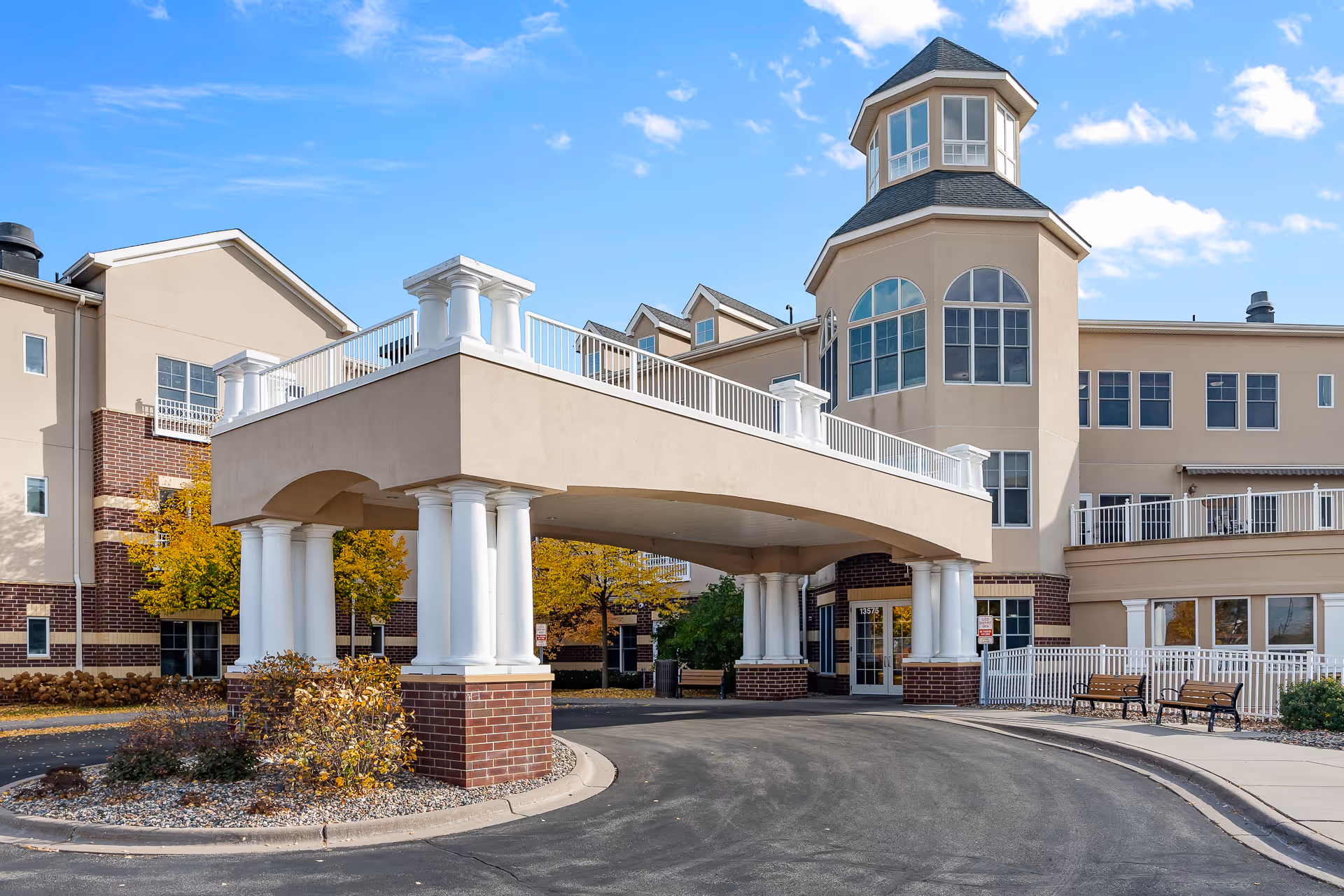 Exterior view of a senior living facility named Boutwells Landing, featuring a covered entrance with white columns and brick accents, a curved driveway, benches, and a multi-story building with large windows and a tower-like structure under a blue sky with scattered clouds.