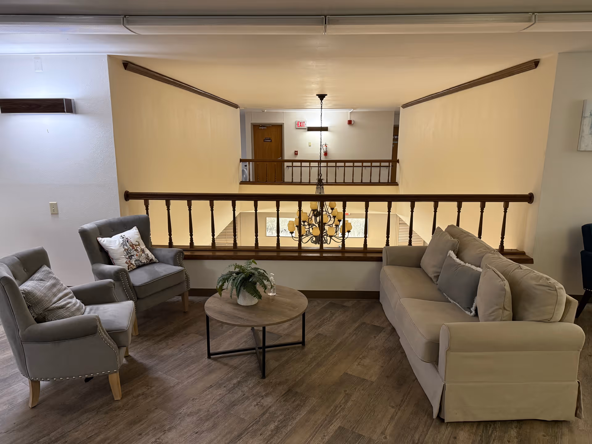 A cozy seating area with a beige sofa and two gray armchairs arranged around a round wooden coffee table with a potted plant. The area overlooks a lower level through a wooden railing, with a chandelier hanging in the open space below. The walls are light-colored and the floor has wood-like flooring.