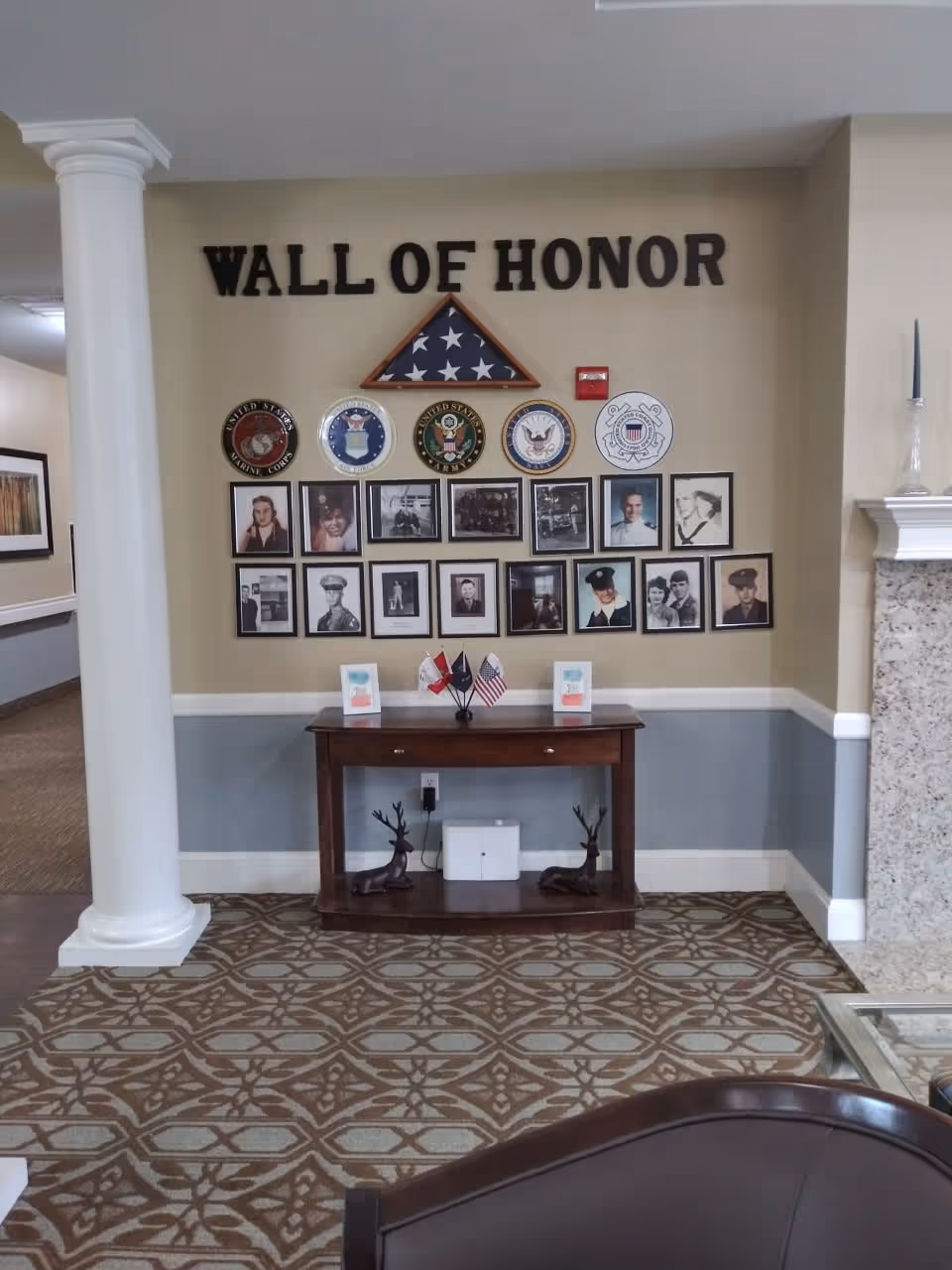 A Wall of Honor display in a senior living facility featuring framed photographs of veterans, military emblems, and a folded American flag in a triangular case mounted on the wall above a wooden table with small flags and decorative items. The area has a patterned carpet, a white column on the left, and a fireplace on the right.