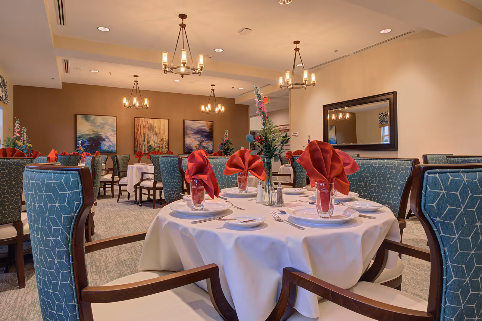 Elegant dining room with round tables set with white linens and red napkins, surrounded by upholstered chairs and chandeliers.