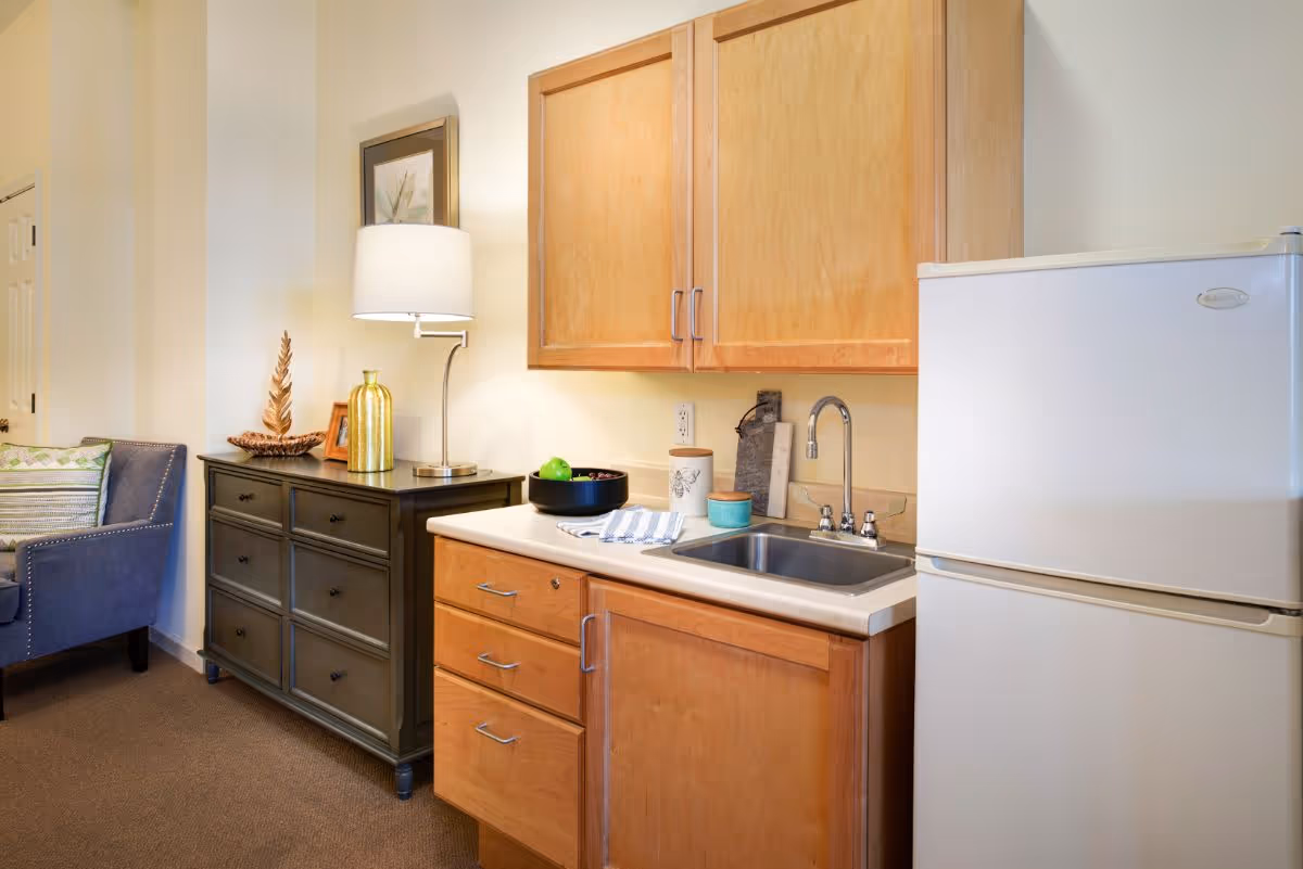 A small kitchenette area with wooden cabinets, a white countertop, a stainless steel sink, and a white refrigerator. Next to the kitchenette is a dark wooden dresser with a table lamp, decorative items, and a framed picture on the wall above it. A blue upholstered chair with patterned cushions is partially visible on the left side.