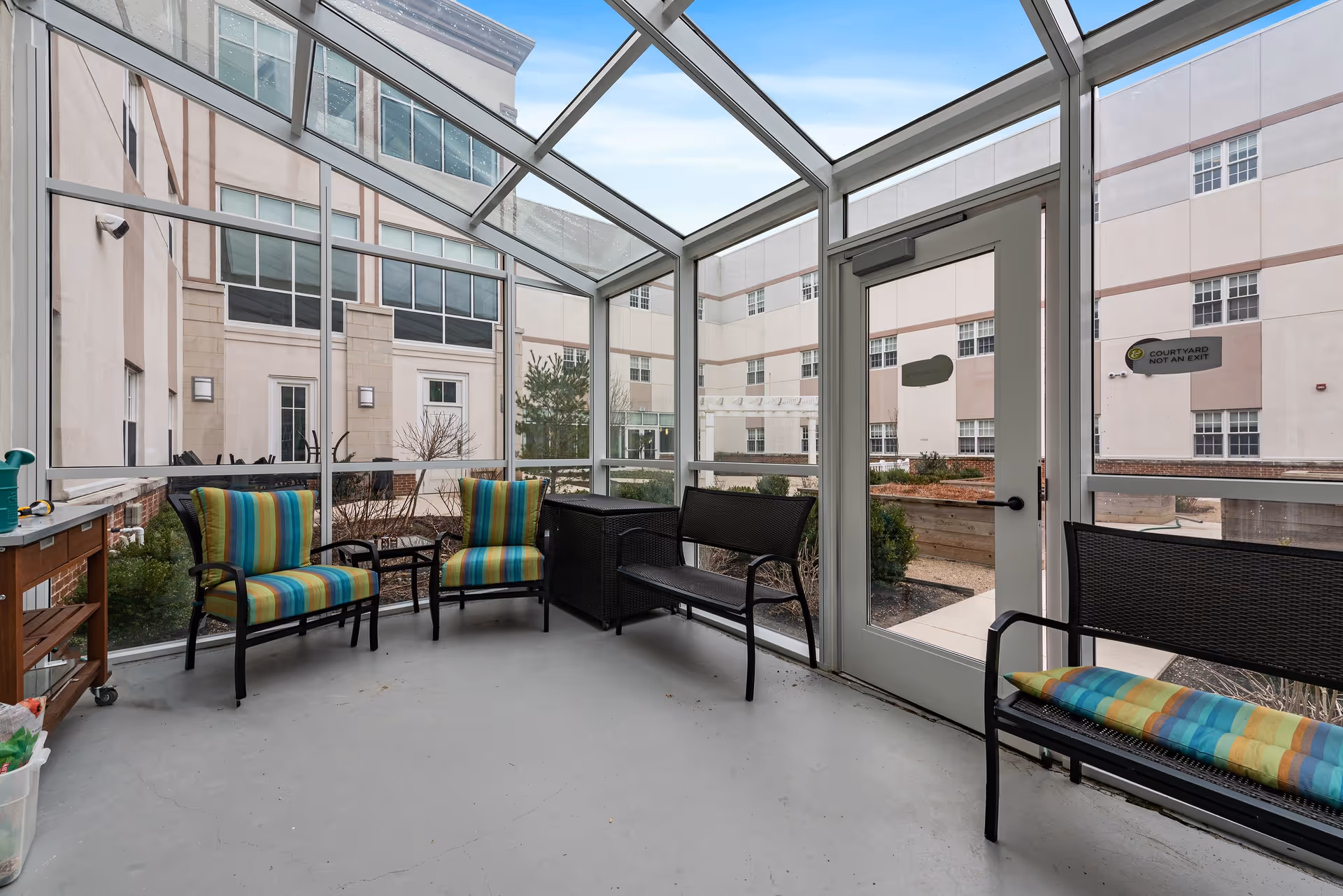 A sunroom or enclosed patio area with glass walls and ceiling, containing two striped cushioned chairs, a bench with a striped cushion, a small table, and a wooden cart. Outside the glass enclosure, there is a courtyard with garden beds and a building with multiple windows.