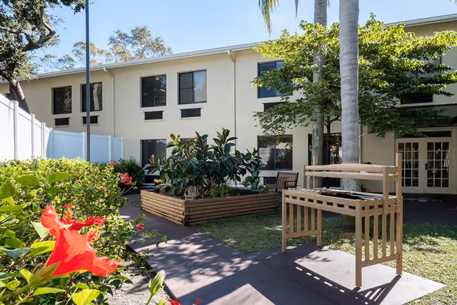 Outdoor garden area at a senior living facility with a raised wooden planter filled with green plants, a wooden bench, palm trees, and a beige two-story building in the background under a clear blue sky.