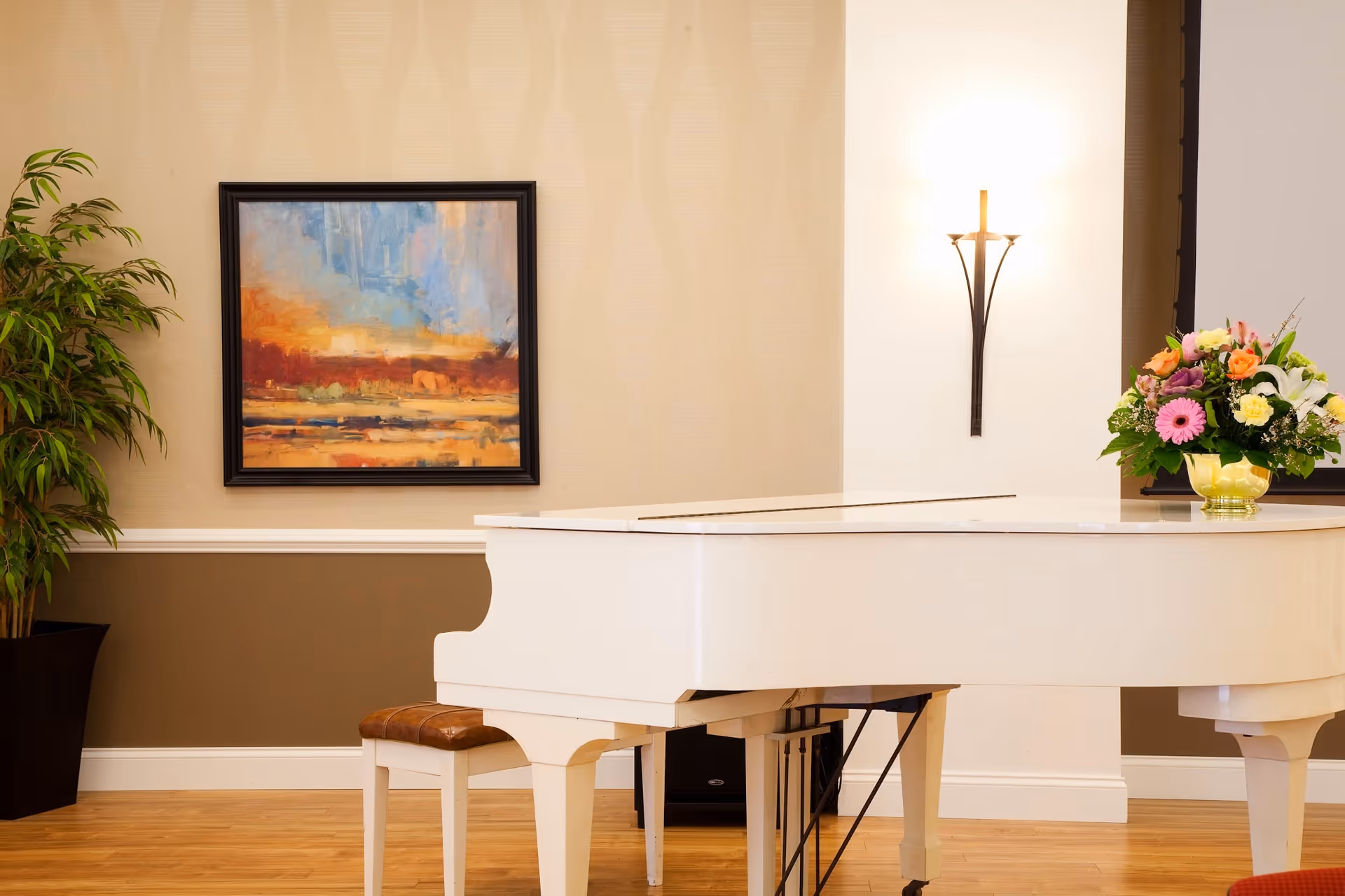 A white grand piano with a brown cushioned bench in a room with wooden flooring. On the piano is a vase with a colorful bouquet of flowers. The wall behind has a framed abstract painting with warm colors and a modern wall sconce light fixture. A tall green plant is placed in a black pot to the left.