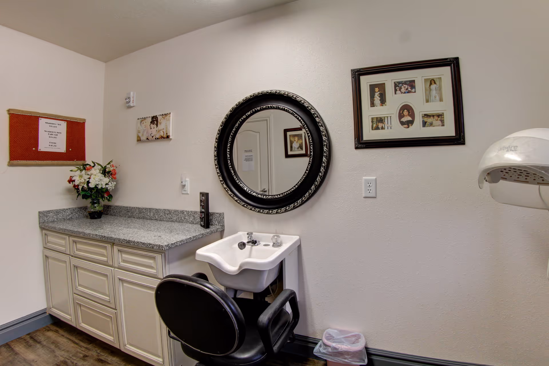 Interior view of a hair salon area in a senior living facility featuring a black salon chair in front of a white sink with a round decorative mirror above it. To the left is a countertop with cabinets underneath and a vase of flowers. On the wall are framed pictures and a red bulletin board. A hair dryer is mounted on the right side of the image.