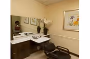 A salon-style station with a mirror, marble countertop, two vases of white flowers, framed art, and a black styling chair in a beige room.