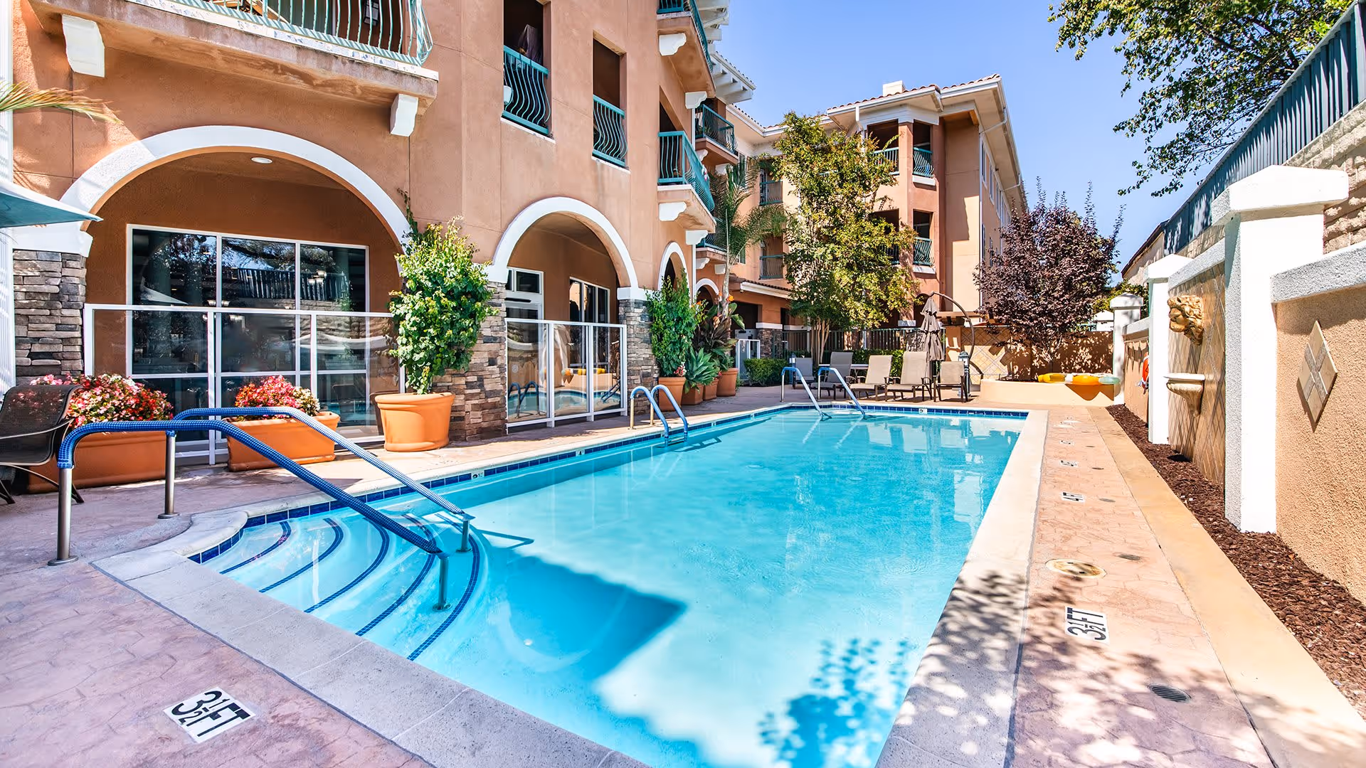 Outdoor swimming pool area at a senior living facility with clear blue water, steps with handrails leading into the pool, surrounded by a patio with potted plants, lounge chairs, and a multi-story building with balconies in the background under a clear blue sky.