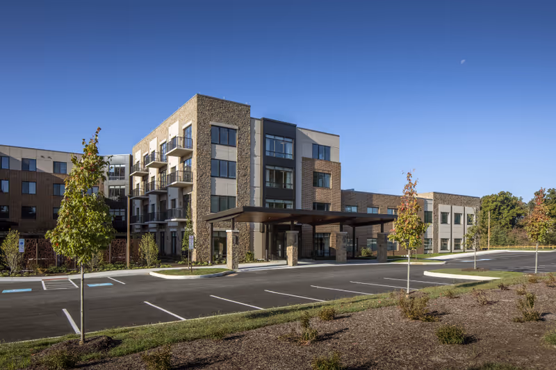 Exterior view of a modern multi-story senior living facility with stone and brick facade, large windows, balconies, a covered entrance, and a parking lot with young trees and landscaping under a clear blue sky.