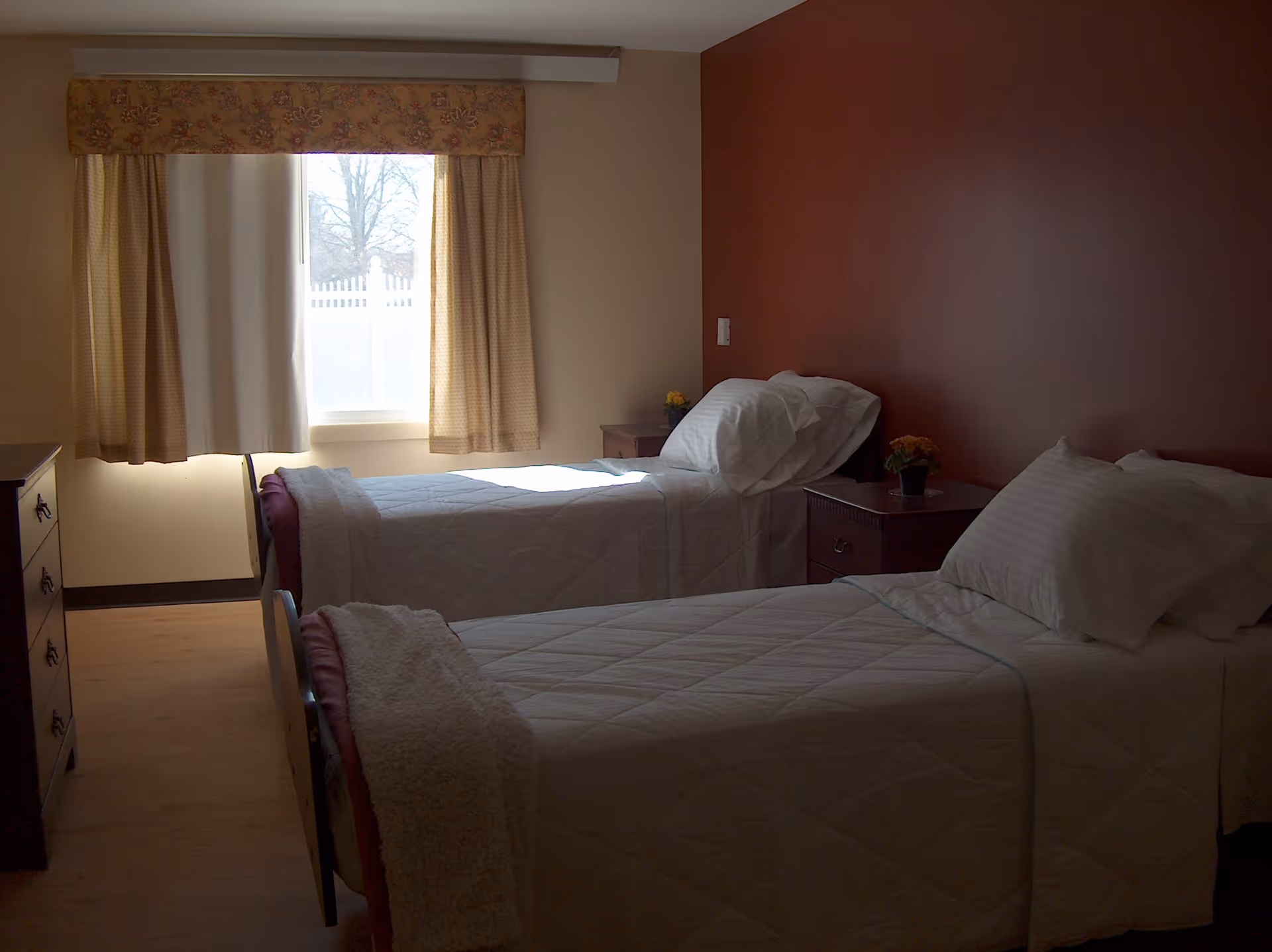 A bedroom with two single beds covered in white quilts and pillows. There is a window with beige curtains letting in natural light. The walls are painted beige and one wall is a dark reddish-brown color. There are two nightstands with small potted plants and a dresser on the left side of the room.