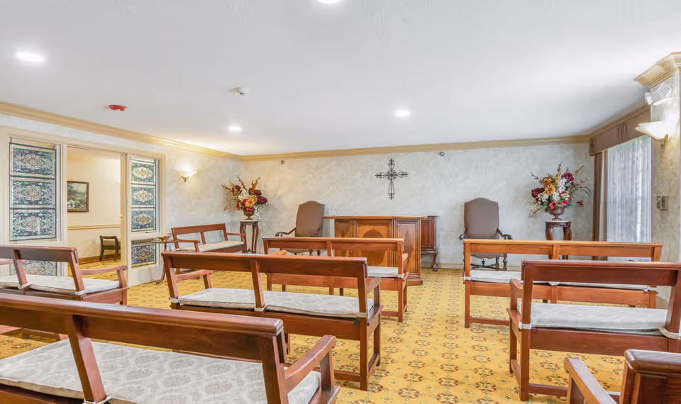 Interior view of a small chapel or meditation room with wooden benches arranged in rows facing a wooden altar. The room has floral arrangements on either side of the altar, a decorative cross on the wall, patterned carpet, and soft lighting.