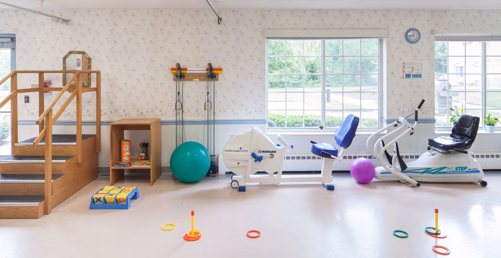 A bright rehabilitation/exercise room with therapy stairs, recumbent exercise bikes, stability balls, resistance bands, and colorful floor rings.
