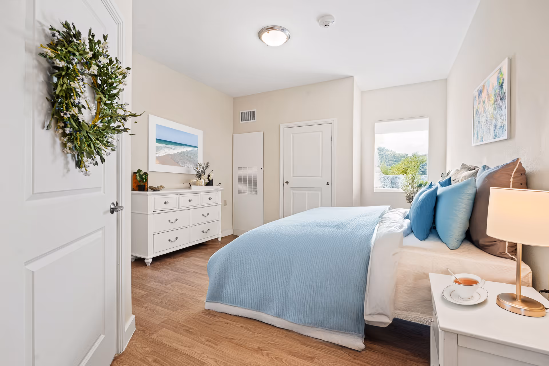 A bright and cozy bedroom with a bed covered in light blue bedding and multiple pillows. There is a white nightstand with a lamp and a cup of tea on it. A white dresser with decorative items and a framed beach picture hangs above it. A wreath is hanging on the door, and a window lets in natural light, showing greenery outside.