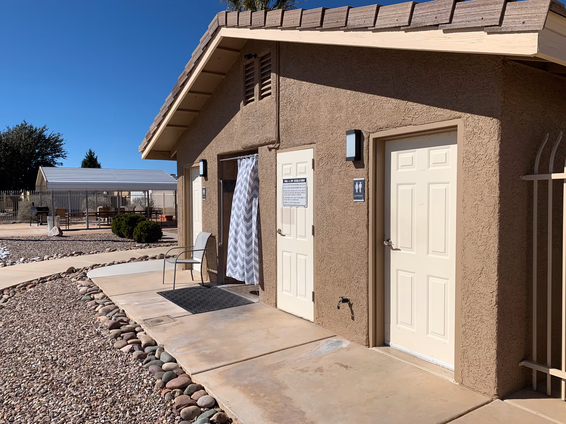 Exterior view of a small building with beige stucco walls and a tiled roof under a clear blue sky. The building has three doors, one with a chevron-patterned curtain, and two solid doors with restroom signs. A metal chair is placed outside near the curtained door. The surrounding area has a concrete walkway, decorative rocks, and some bushes. In the background, there is a covered seating area with tables and chairs.