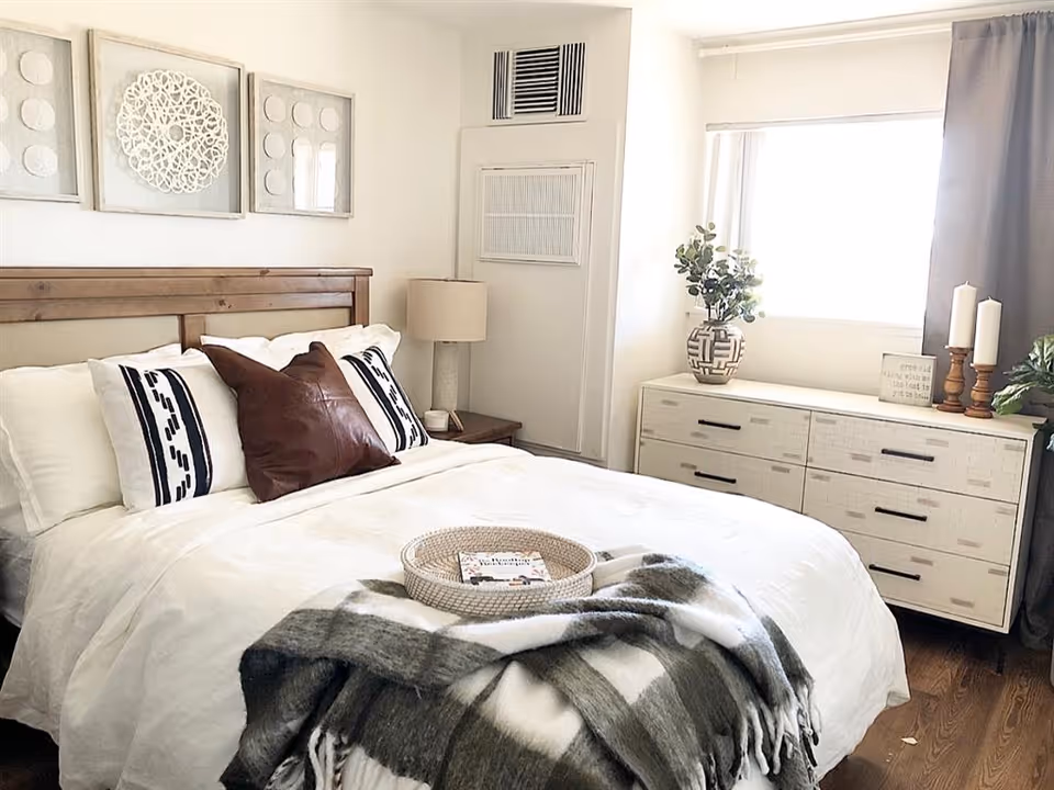 Bright, styled bedroom with a white bed topped with pillows and a plaid throw, a wooden headboard, bedside lamp, and a dresser beneath a window.