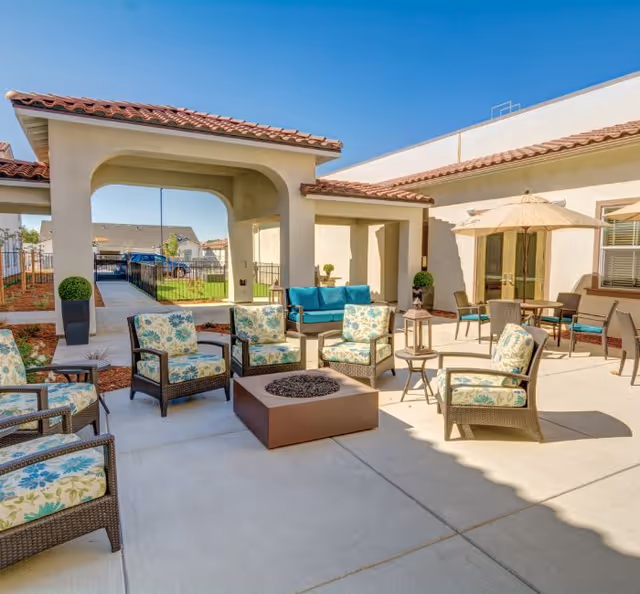 Outdoor patio area at The Village at Heritage Park with cushioned wicker chairs arranged around a square fire pit. There is a covered seating area with a blue cushioned bench, a table with chairs and an umbrella, and potted plants. The building has a light stucco exterior with a red tile roof.