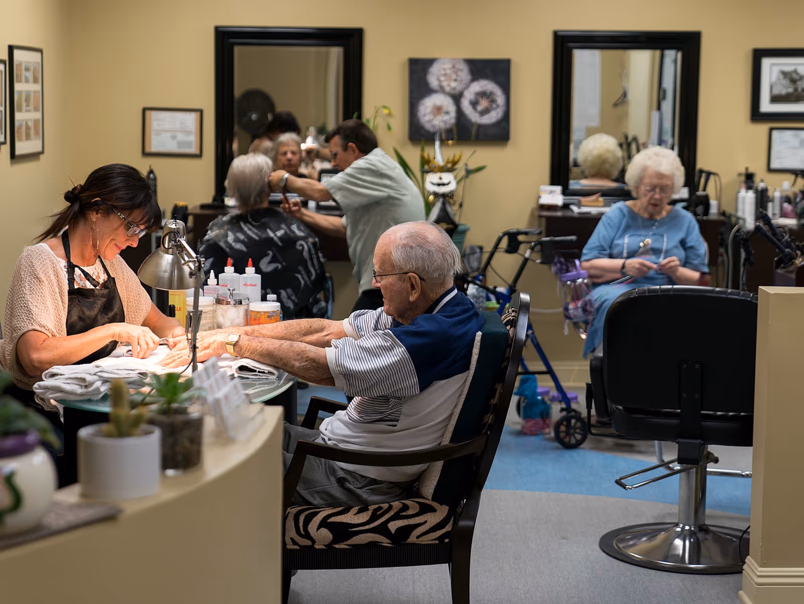 An elderly man is getting a manicure from a woman in a salon area, while in the background an elderly woman is seated near a walker and a man is cutting another person's hair. The room has mirrors, framed pictures, and salon equipment.