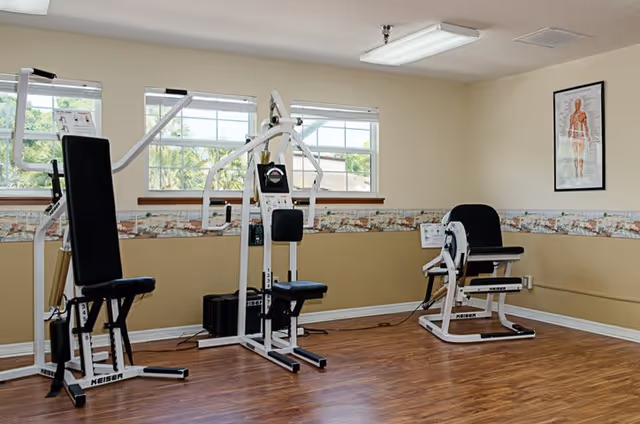 A small exercise room with two pieces of white and black Keiser fitness equipment on a wooden floor. The room has beige walls with a decorative border and three windows letting in natural light. A framed anatomical poster is hung on the wall.