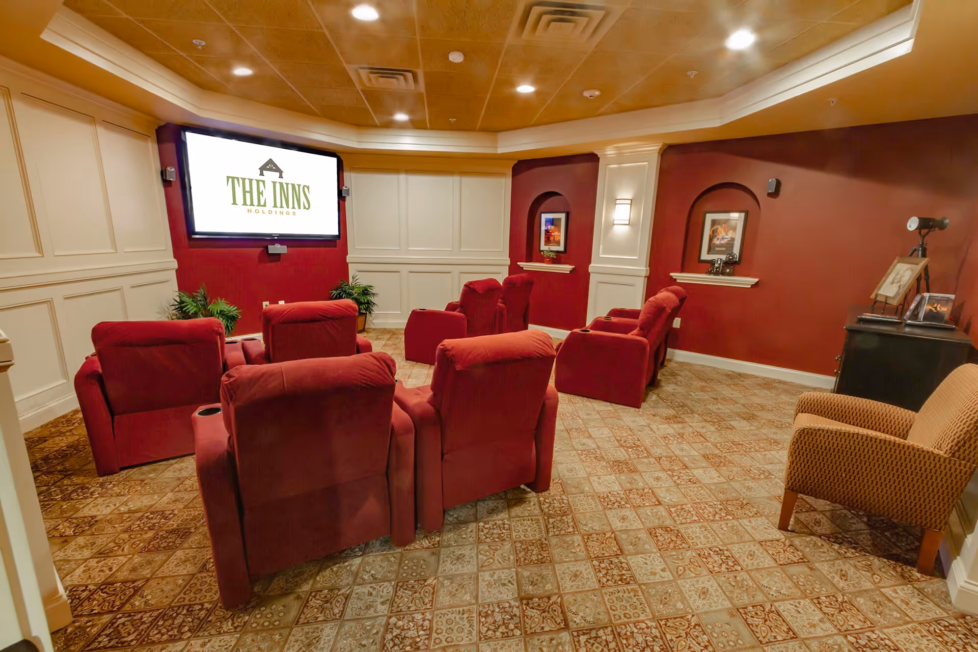 A cozy media room with eight red recliner chairs arranged in two rows facing a large wall-mounted screen displaying 'THE INNS HOLDINGS'. The room has patterned tile flooring, cream-colored wainscoting on the walls, and a warm wooden ceiling. There are framed pictures on the red walls and a beige armchair to the right.