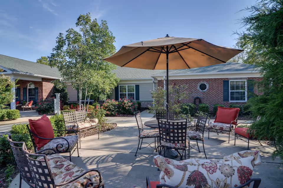 Outdoor patio area at Hickory Gardens with metal chairs and benches featuring floral cushions and red pillows arranged around a round table with a large beige umbrella. The patio is surrounded by greenery, bushes, and trees, with a brick building in the background under a clear blue sky.