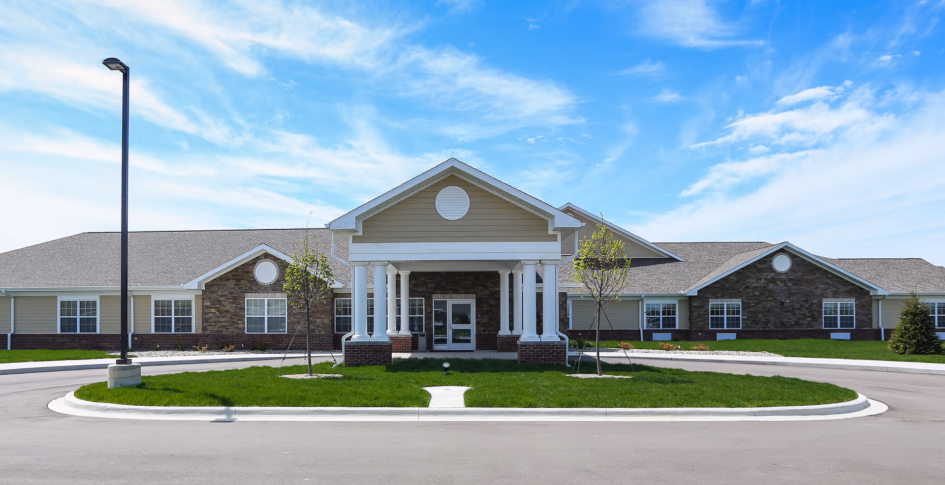 Front entrance of a single-story senior living building with white columns, gabled roof, stone and siding facade, circular driveway and manicured lawn under a blue sky.