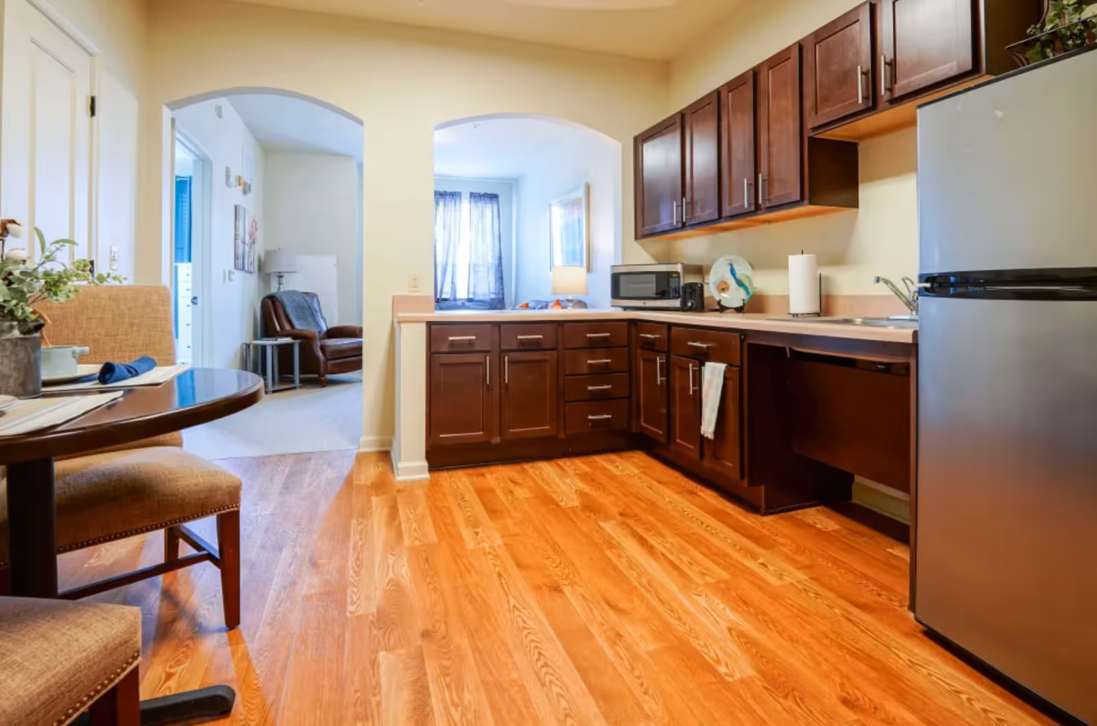 A kitchen area with wooden cabinets, a countertop with a microwave, paper towel holder, and decorative plate. There is a stainless steel refrigerator on the right and a small dining table with two chairs on the left. The kitchen opens into a living area with a leather armchair and a window with sheer curtains.