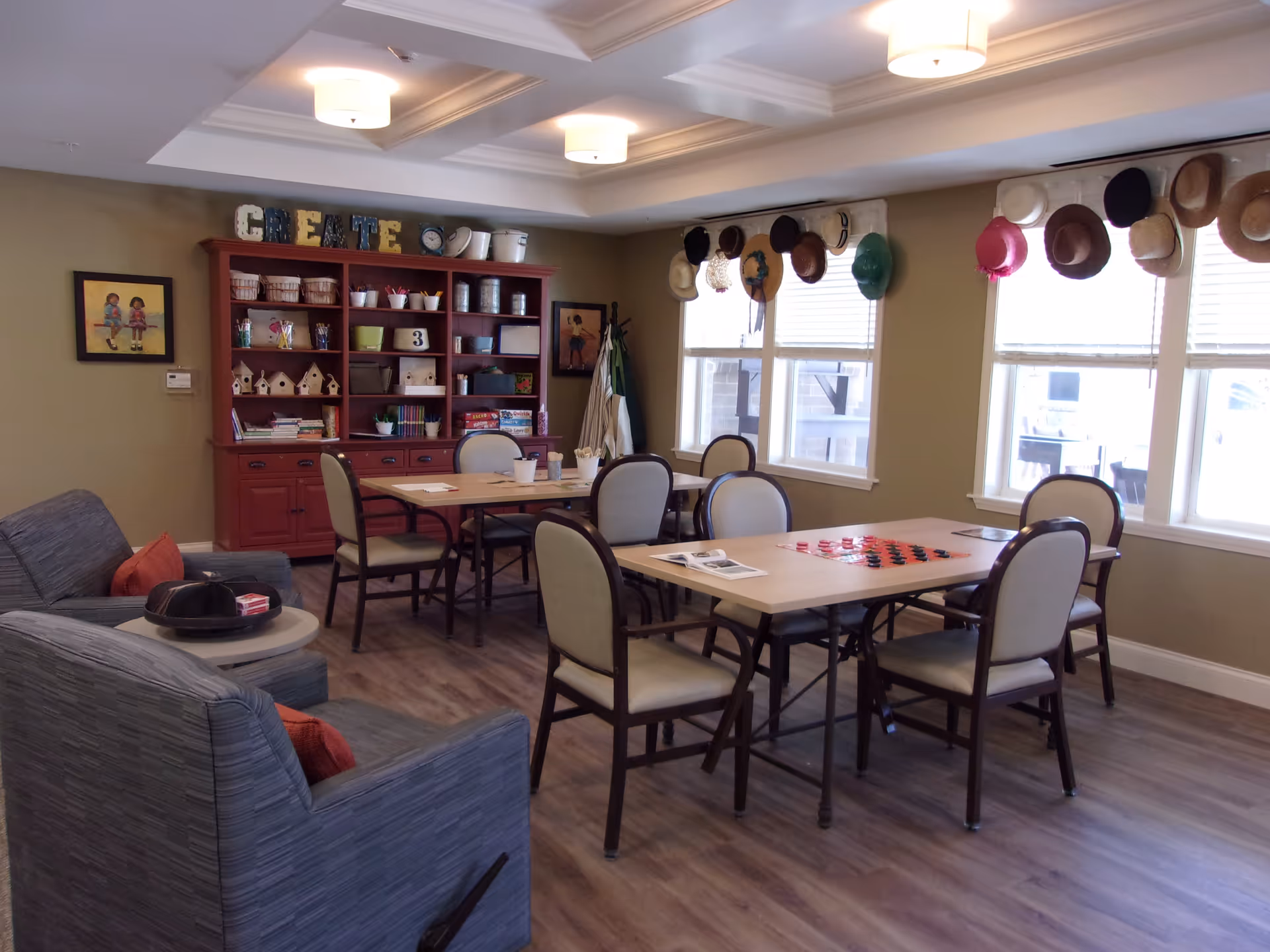 A cozy activity room with two tables and several chairs arranged around them. One table has a checkers game set up, and the other has some papers and cups. There are two comfortable armchairs with orange cushions in the foreground. A red cabinet filled with craft supplies and games is against the back wall, with the word 'CREATE' displayed on top. Several hats hang on hooks above the windows on the right side of the room. The room has wood flooring and soft lighting from ceiling fixtures.