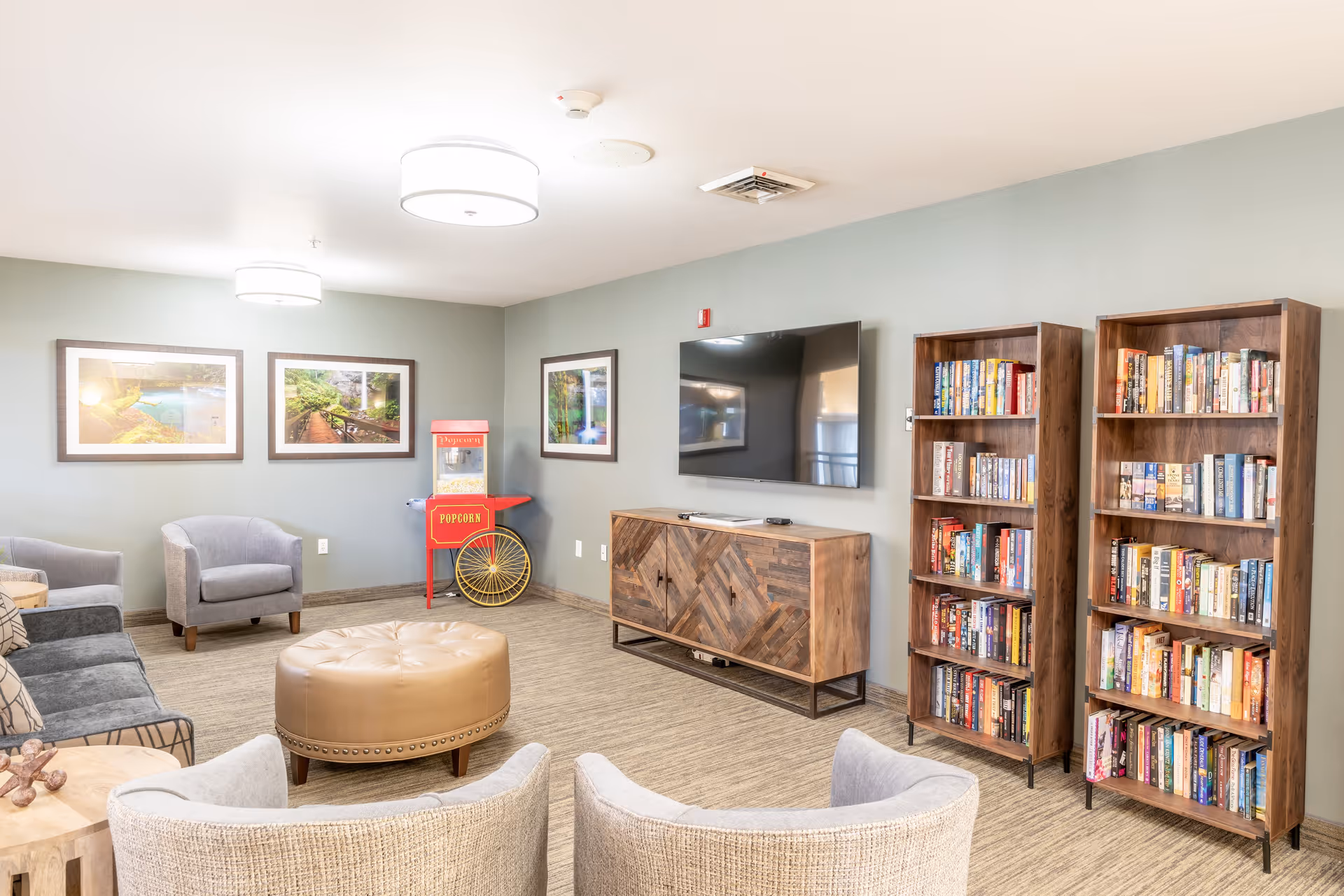 A cozy senior living common area with comfortable seating including armchairs and a sofa, a round leather ottoman in the center, two wooden bookshelves filled with books, a wooden cabinet with a flat-screen TV mounted above it, framed nature photographs on the walls, and a red popcorn machine in the corner.