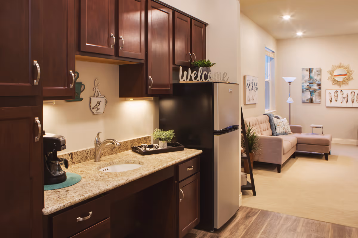 Interior view of a senior living facility showing a kitchenette with dark wood cabinets, a granite countertop with a sink, a coffee maker, and a small refrigerator. The kitchenette opens into a living area with a beige sectional sofa, a floor lamp, wall art, and a decorative mirror.