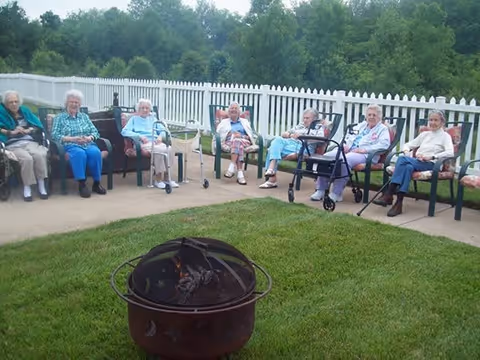 Seven elderly women sitting in chairs arranged in a semicircle on a patio area with a white picket fence and green trees in the background. In the foreground, there is a fire pit on a grassy area.