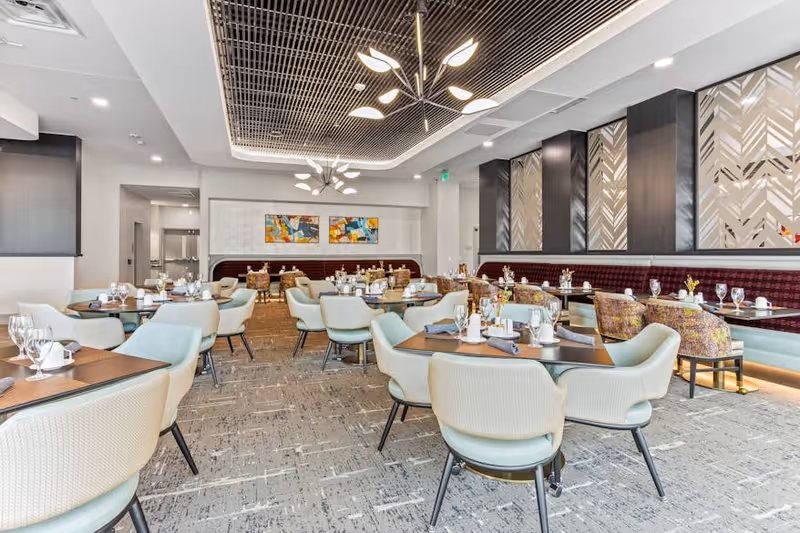 Bright modern dining room with multiple tables and upholstered chairs set with glassware and place settings.