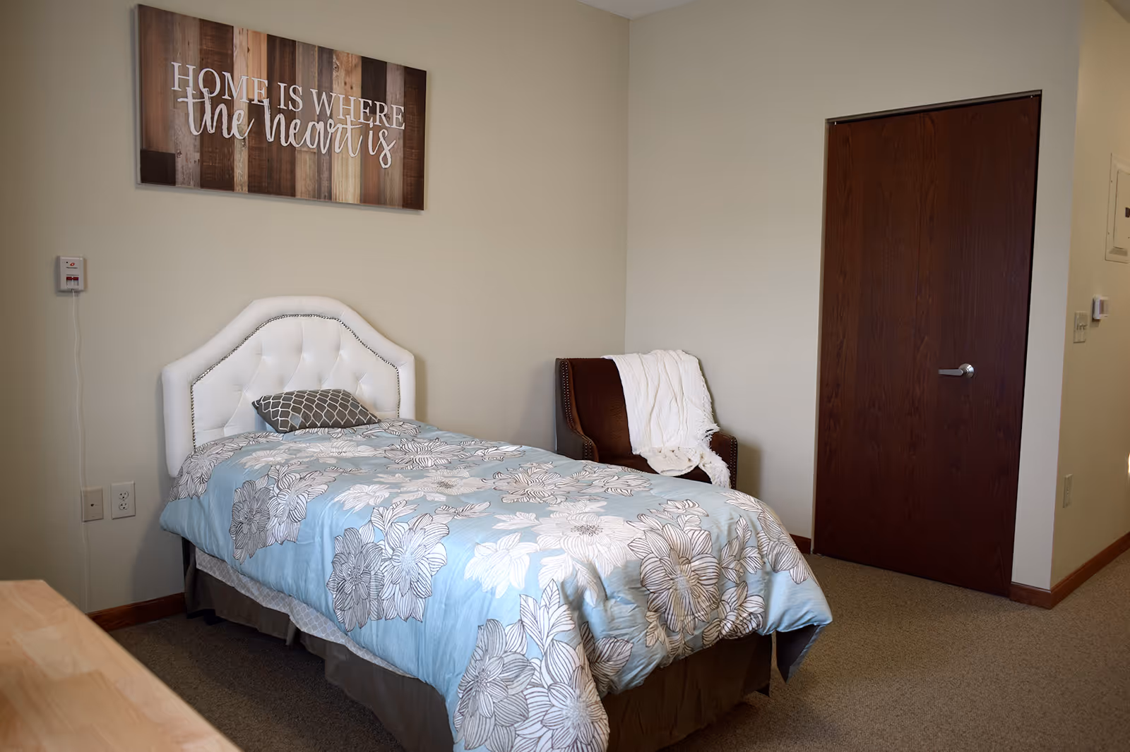 A neatly made single bed with a white tufted headboard and a blue floral comforter in a simple bedroom. A brown armchair with a white throw blanket is placed next to the bed. A wooden door is visible on the right wall, and a decorative sign above the bed reads 'Home is where the heart is.'