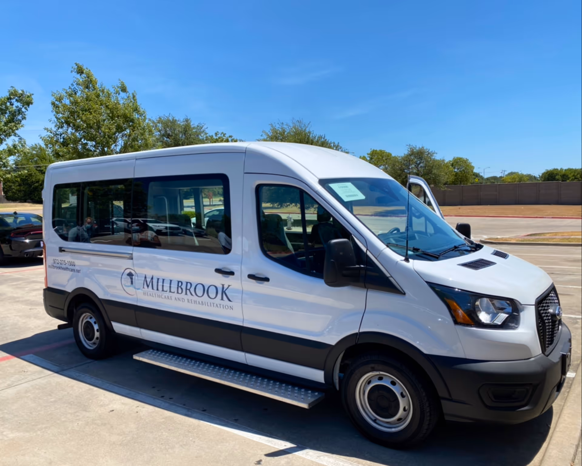 White shuttle van parked outdoors on a sunny day with the logo and name Millbrook Healthcare and Rehabilitation Center on the side.
