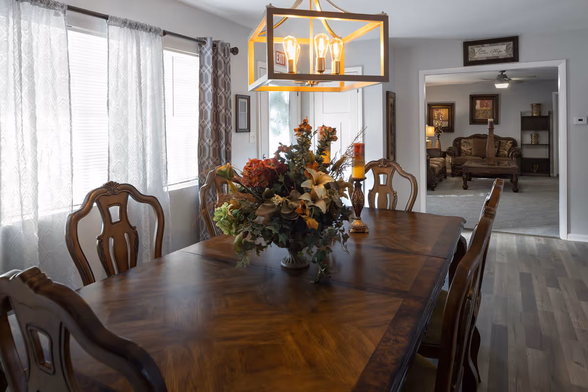 Wooden dining table with a floral centerpiece and chairs in a bright dining room opening to a living room.